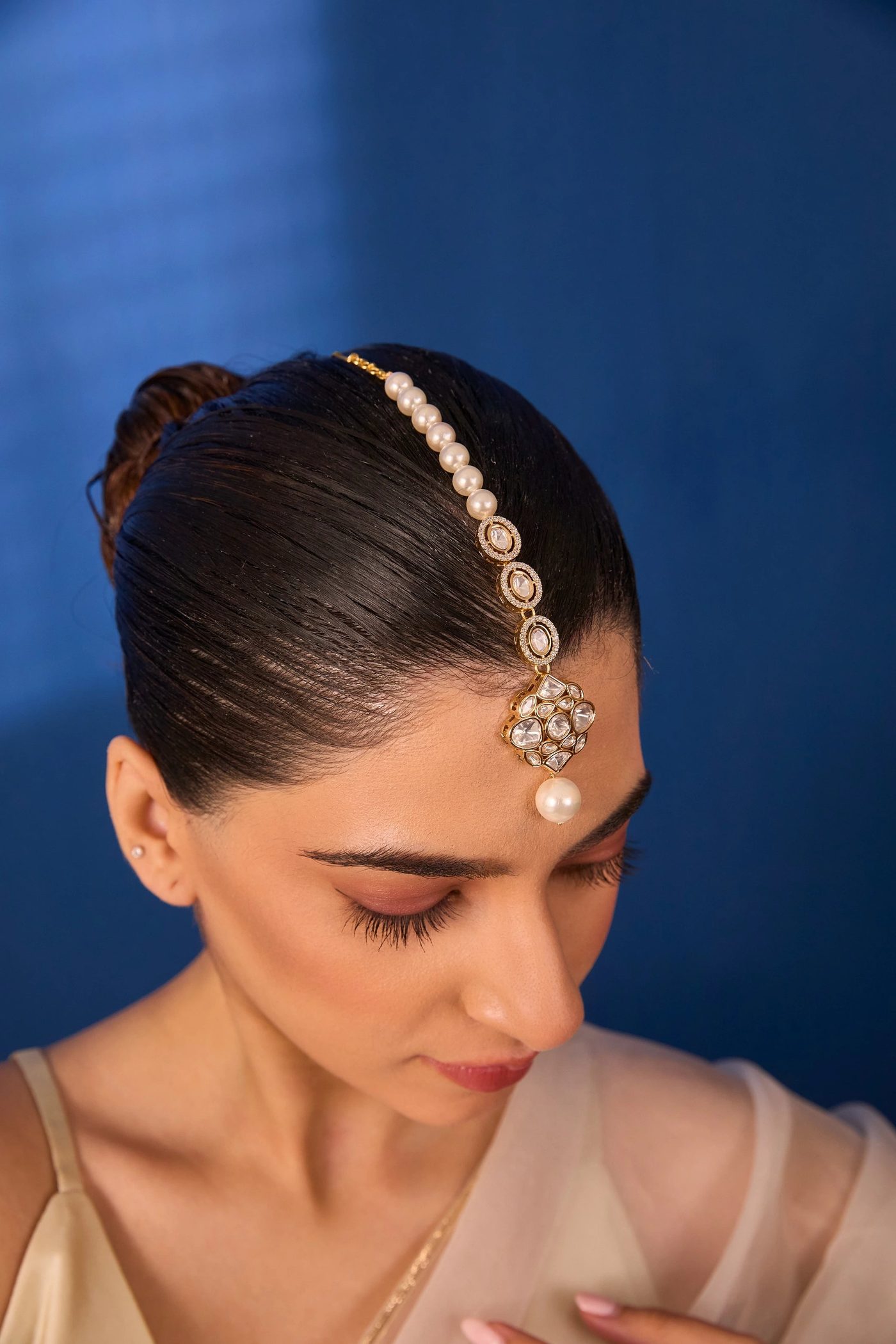 A pearl and polki maang tikka worn with a sleek bun against a lapis-blue wall