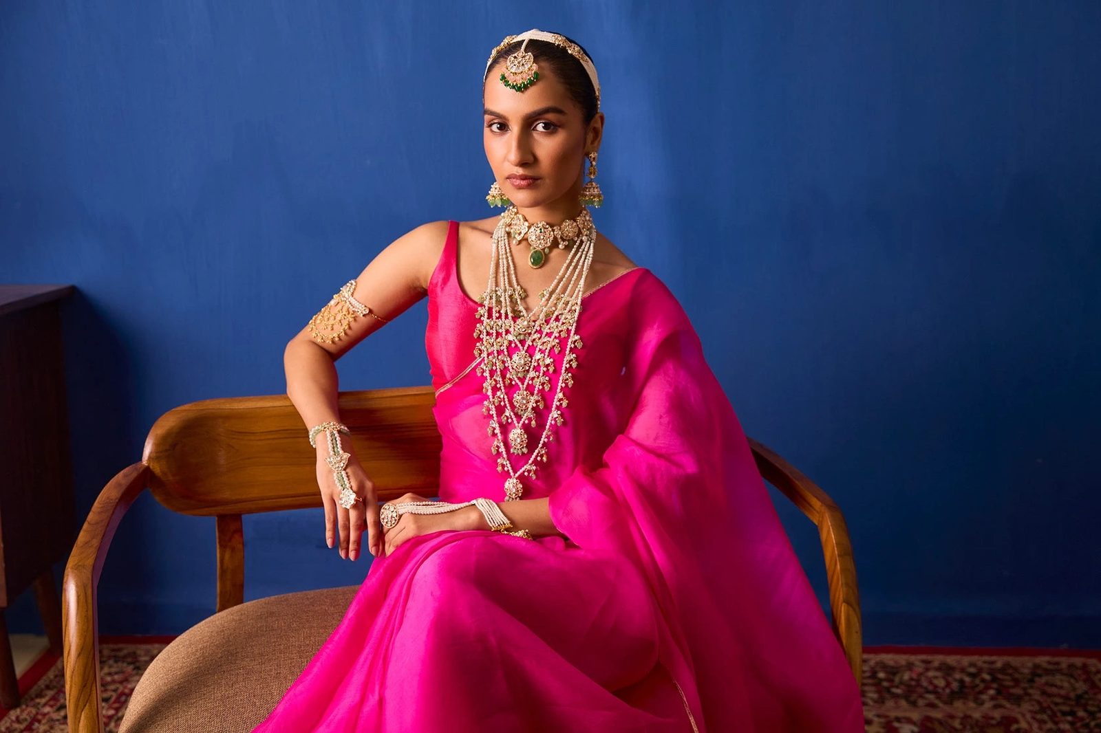 A woman in a pink silk saree against a lapis-blue wall, wearing a polki choker, layered pearl necklaces, maang tikka and hand jewellery